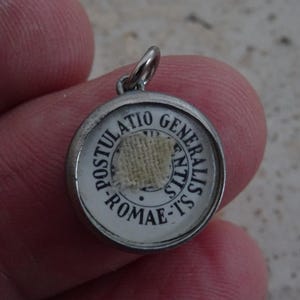 Pode incluir: Pingente redondo prateado com fundo branco e texto preto. O texto diz "POSTULATIO GENERALIS ROMAE-TS". O centro apresenta um pequeno pedaço quadrado de tecido bege. O pingente tem uma pequena argola na parte superior.