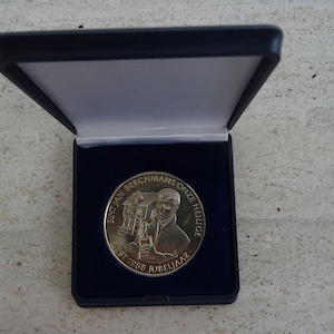 May include: A silver coin in a blue velvet box. The coin features a portrait of a man and the text "Sint Jan Berchmans Onze Heilige" and "Diest 1938 Jubilejaar".