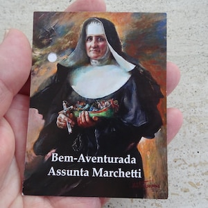 May include: A religious card featuring a portrait of a nun in a black habit and white wimple. She holds a cross and a small boat with figures. The card has the text "Bem-Aventurada Assunta Marchetti".