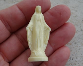 以下が含まれることがあります： 両手を広げた、ローブを着た人物の小さなクリーム色の置物。像は小さな台座の上に立っています。この人物は穏やかな表情をしており、おそらく宗教的なアイコンです。この置物は滑らかな、おそらくプラスチック素材で作られています。