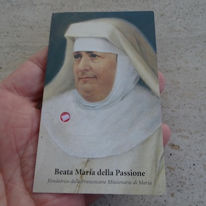 Könnte beinhalten: Eine rechteckige Gebetskarte mit dem Porträt einer Frau in weißem Habit und Schleier. Der Text am unteren Rand lautet Beata Maria della Passione, Fondatrice delle Francescane Missionarie di Maria, versehen mit einem kleinen roten Aufkleber.