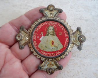 May include: A vintage metal religious cross with a red and gold circular medallion featuring a depiction of Jesus Christ with the words "COR JESV SACRATISSIMVM MISERERE NOBIS MONTMARTE" around the image.