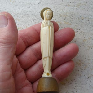 May include: A small ivory figurine of a woman in a long robe with her hands clasped in prayer. The figurine is standing on a wooden base.