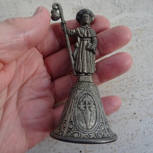May include: A silver-coloured figurine of a pilgrim holding a staff and satchel, atop a bell-shaped base with an ornate design. The figure is detailed, with a hat and robe. The base features a cross symbol.