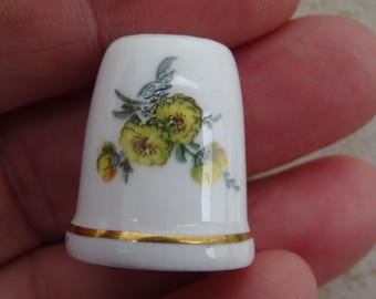 May include: A white ceramic thimble with a gold trim and a floral design featuring yellow flowers and green leaves.
