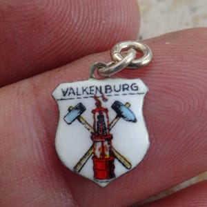 Peut inclure: Un petit pendentif en forme de bouclier avec le mot "VALKENBURG" en haut. Le pendentif présente un fond blanc avec un dessin de marteaux croisés et une structure minière en rouge, jaune et bleu. Le pendentif est attaché à un anneau argenté.
