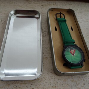 May include: A wristwatch with a green band and a black watch face. The watch face has a green background with a portrait. The watch is in a silver rectangular tin.