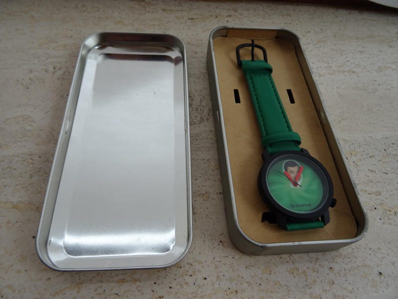 May include: A wristwatch with a green band and a black watch face. The watch face has a green background with a portrait. The watch is in a silver rectangular tin.