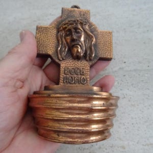 Puede incluir: Una pequeña cruz de metal dorado con una representación del rostro de Jesucristo y la inscripción "Ecce Homo" en la base.