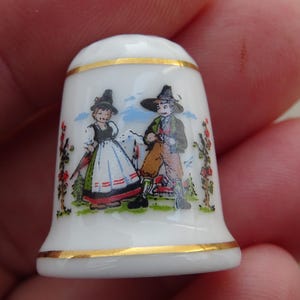 Może przedstawiać: Biały naparstek z porcelany z ręcznie malowaną ilustracją przedstawiającą dwie postacie w tradycyjnych strojach na tle krajobrazu. Naparstek jest ozdobiony złotymi paskami na górnej i dolnej krawędzi, które obramowują kolorową scenę z parą.