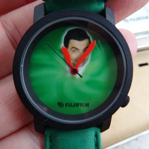 May include: Black wristwatch with a green face featuring a portrait of Mr. Bean. The watch has red hands and a green leather strap. The brand name "FUJIFILM" is printed on the watch face.