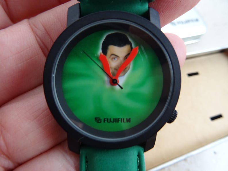 May include: Black wristwatch with a green face featuring a portrait of Mr. Bean. The watch has red hands and a green leather strap. The brand name "FUJIFILM" is printed on the watch face.