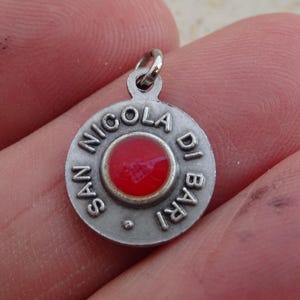 Pode incluir: Um pequeno pingente redondo de prata com as palavras "SAN NICOLA DI BARI" circundando um círculo vermelho central. O pingente tem um pequeno laço na parte superior para prender. O pingente está na mão de uma pessoa.