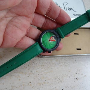 May include: A green wristwatch with a black face and green leather strap. The watch face features a cartoon character on a green background. The watch is held in a hand, and is next to a metal tin.