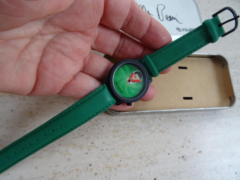 May include: A green wristwatch with a black face and green leather strap. The watch face features a cartoon character on a green background. The watch is held in a hand, and is next to a metal tin.