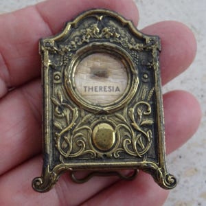 Puede incluir: Un pequeño relicario rectangular de latón de estilo antiguo con detalles en relieve de flores y enredaderas. El centro contiene una ventana circular de cristal que encierra un fondo de tela con el nombre THERESIA impreso en letras mayúsculas negras.