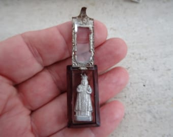 May include: A small, silver-colored religious pendant with a clear glass front and a brown metal back. The pendant features a depiction of a religious figure in a white robe.