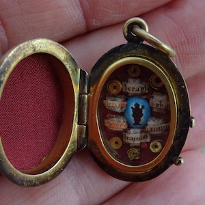 May include: An oval brass locket with a hinged lid, featuring a red fabric interior. The right side contains a central blue oval stone with a black silhouette, surrounded by small gold-toned metal rings and white paper strips with text.