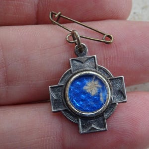 Pode incluir: Um pingente de cruz de prata com um centro de esmalte azul que apresenta uma flor branca. O pingente está preso a um alfinete de segurança.