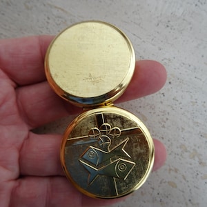 Könnte beinhalten: Ein goldfarbener, runder Kompaktspiegel, geöffnet, um ein detailliertes Design zu zeigen. Die untere Hälfte zeigt ein geprägtes Bild von zwei Fischen und anderen geometrischen Formen. Die obere Hälfte ist eine schlichte, reflektierende Oberfläche.