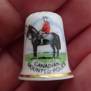 May include: White porcelain thimble with a gold trim. It features a colorful illustration of a mounted Canadian police officer riding a black horse. The text "Canadian Mounted Police" is printed on the thimble.