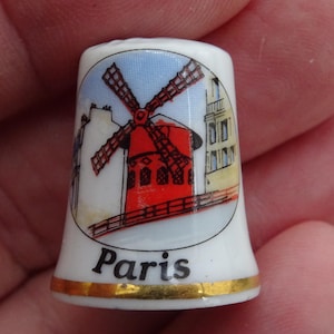 Puede incluir: Dedal de cerámica blanca con un diseño de molino de viento rojo, aspas negras y edificios, sobre un cielo azul. La palabra "París" está impresa debajo, con una banda dorada en la base. Un artículo de recuerdo.