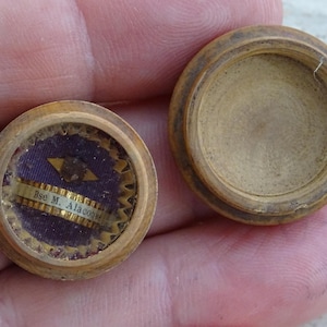 Pode incluir: Um pequeno recipiente de madeira aberto com formato circular. O interior do recipiente apresenta um tecido roxo com uma faixa dourada com o texto "Bse M. Alacon". A tampa está separada, revelando o interior do recipiente.