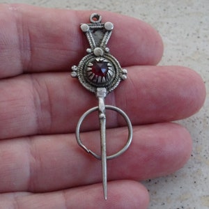 May include: A silver brooch with a red gemstone in the center. The brooch has a decorative design and a long pin.