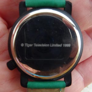 May include: A black wristwatch with a green strap. The watch face is black with a silver rim. The text "Tiger Television Limited 1999" is visible on the watch face.