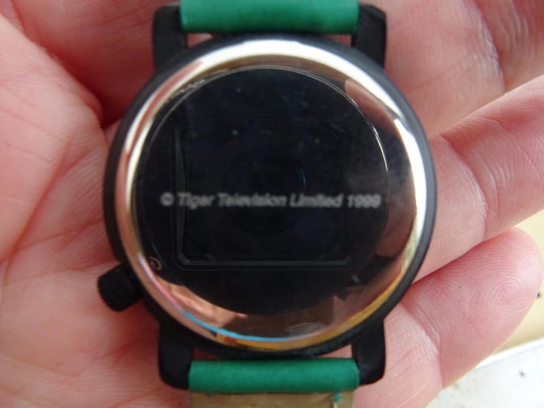 May include: A black wristwatch with a green strap. The watch face is black with a silver rim. The text "Tiger Television Limited 1999" is visible on the watch face.