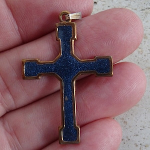 Pode incluir: Um pingente de cruz em tom dourado com acabamento em esmalte azul. O esmalte tem um aspecto texturizado e salpicado.