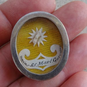 Può includere: Un ciondolo rotondo argentato con un fronte in vetro. Il ciondolo contiene un tessuto giallo con una stella bianca e il testo "unita S.S. Mart. Gorcu" in bianco.
