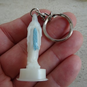 May include: White plastic keychain featuring a small figurine of a woman in a blue robe. The keychain has a silver metal ring.