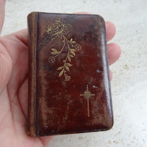 Puede incluir: Un libro pequeño y rectangular encuadernado en cuero marrón oscuro con una superficie desgastada y texturizada. La portada presenta un diseño floral dorado en relieve en la esquina superior izquierda y una pequeña cruz dorada punteada.