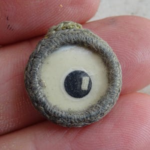 Pode incluir: Um botão pequeno, redondo, cinza e branco com um design de olho preto e branco. O botão é feito de tecido e tem um centro branco com um olho preto no centro. O olho tem uma linha branca que o atravessa.