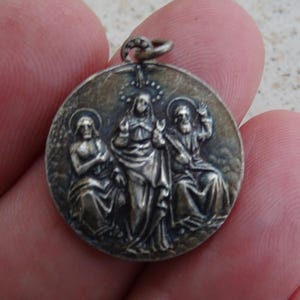 May include: A round, silver-toned pendant featuring three figures in relief. The central figure is flanked by two others, all with halos. The pendant has a loop at the top for a chain.