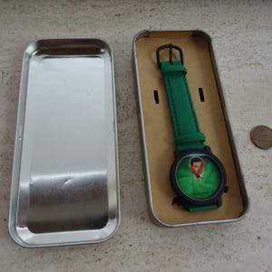 May include: A green wristwatch with a black case and a green strap, presented in a silver metal box. The watch face features a cartoon image. A penny is next to the box.