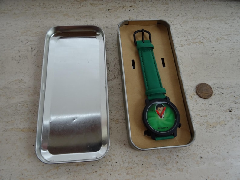 May include: A green wristwatch with a black case and a green strap, presented in a silver metal box. The watch face features a cartoon image. A penny is next to the box.