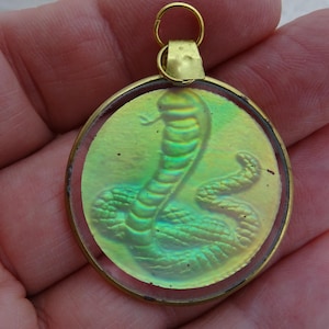 Pode incluir: Um pendente circular com um relevo de serpente iridescente em tons de verde e dourado, inserido numa fina moldura de metal dourado. O pendente inclui um pequeno anel dourado no topo para fixação, apresentando uma superfície brilhante e reflexiva.