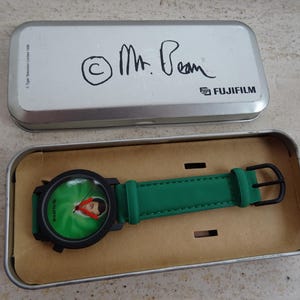 May include: A Mr. Bean wristwatch in a silver Fujifilm tin. The watch has a black case and a green band. The watch face features a green background with a picture of Mr. Bean.