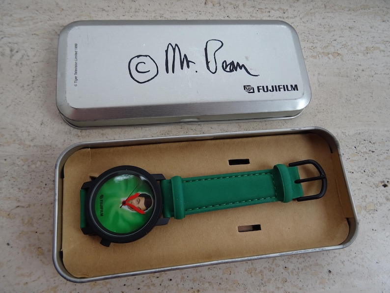 May include: A Mr. Bean wristwatch in a silver Fujifilm tin. The watch has a black case and a green band. The watch face features a green background with a picture of Mr. Bean.
