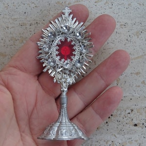 May include: A silver metal religious monstrance with a red glass center and clear rhinestones. The monstrance is on a stand with a decorative base.