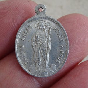 Puede incluir: Colgante ovalado de plata con una figura religiosa sosteniendo un cáliz y un báculo. El colgante tiene las palabras "SAINTE ADELLE" y "PRIEZ P. NOUS" inscritas alrededor de los bordes. Un pequeño lazo en la parte superior para colgar.