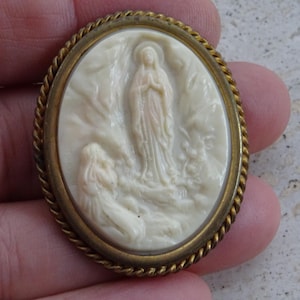 May include: Oval-shaped religious brooch with a cream-colored relief depicting a standing figure and a kneeling figure. The brooch has a gold-colored rope-like border. The figures are set against a textured background.