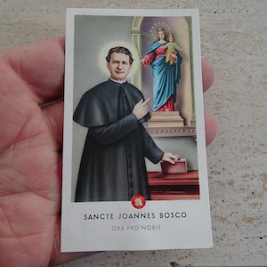 Könnte beinhalten: Religiöse Karte mit einer Illustration eines Mannes in schwarzer Robe mit Heiligenschein, der zeigt. Im Hintergrund eine Darstellung der Jungfrau Maria und des Kindes. Der Text unten lautet "SANCTE JOANNES BOSCO ORA PRO NOBIS."