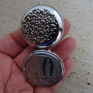 May include: A silver-colored, hinged religious pendant. The top half has a textured, bubble-like design. The bottom half features a detailed relief of a religious scene with a figure in a teal-colored alcove.