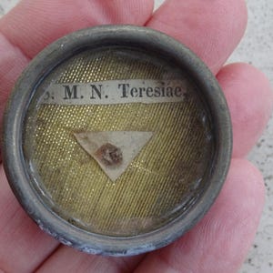 Pode incluir: Uma pequena relíquia religiosa redonda com uma borda metálica. O interior apresenta um tecido dourado, um elemento triangular de papel e uma tira de papel com o texto ". M. N. Teresiae."