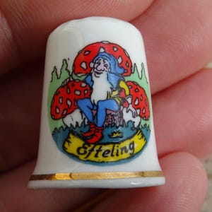May include: White porcelain thimble with a colorful illustration of a gnome sitting on a mushroom. The gnome is wearing a blue hat and a red and green outfit. The thimble has a gold rim and the word "Efteling" written on a yellow banner.