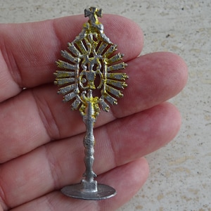 May include: A small, ornate silver-colored religious item with gold accents. The item has a circular base, a slender stem, and a decorative top with a cross. The design features intricate details and a sunburst-like pattern.