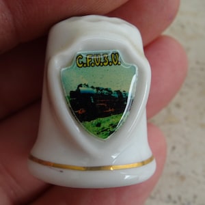 Könnte beinhalten: Weißer Keramik-Fingerhut mit einem goldenen Band am Boden. Der Fingerhut hat ein schildförmiges Emblem mit dem Text "C.F.V.3.U." und ein Bild eines Zuges auf einem grünen Feld.